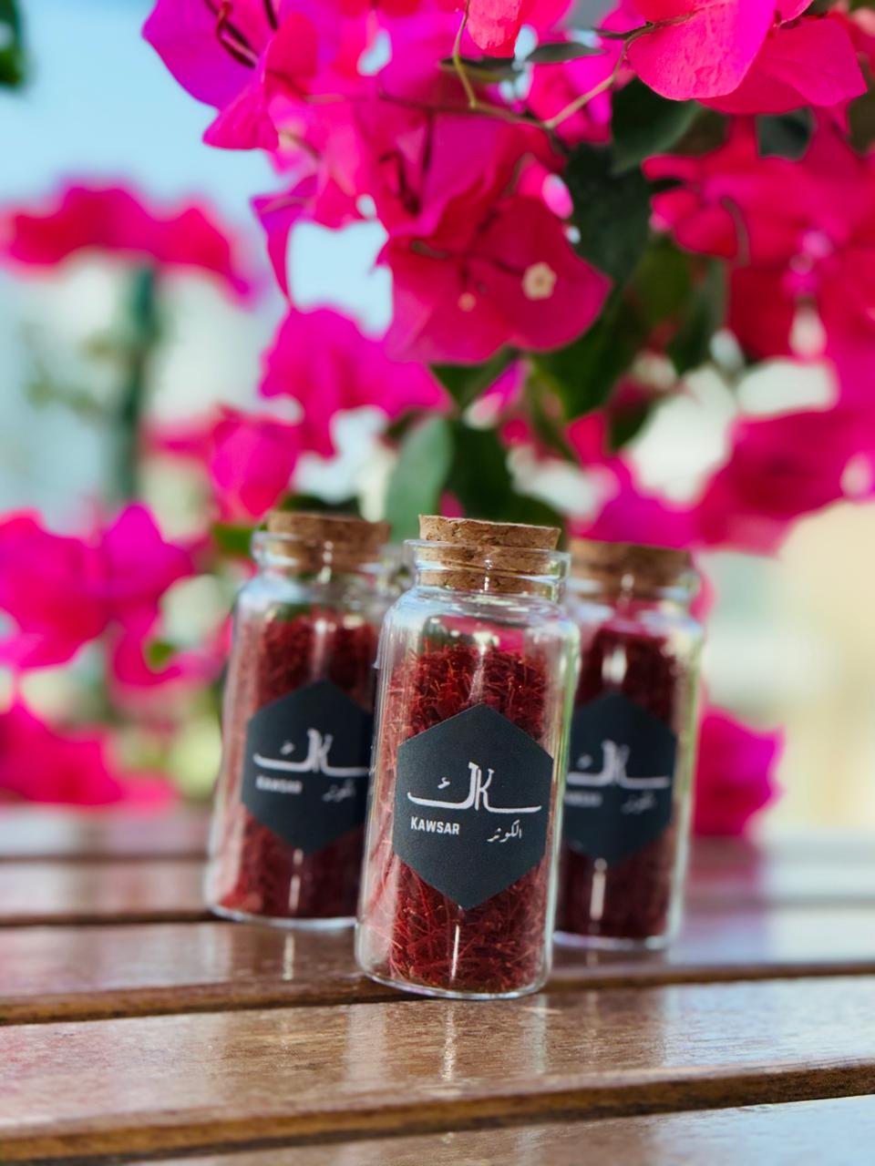 Glass jars of premium Afghan saffron labeled Kawsar, with vibrant pink flowers in the background